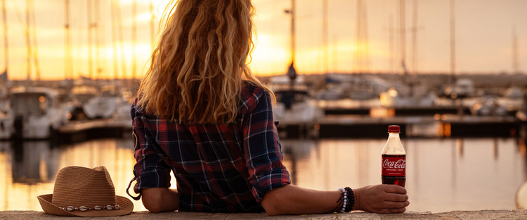 Eine Frau, die mit einer Flasche Coca-Cola in der Hand beim Sonnenuntergang am Hafen Boote betrachtet