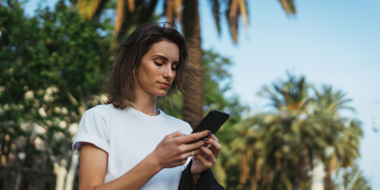 A woman standing outside near palm trees uses an iPhone to engage with a personalized customer experience.