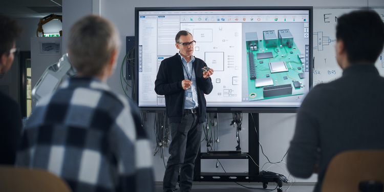 Engineer presenting a 3D circuit board design on a large screen to a group of students in a classroom or lab setting.