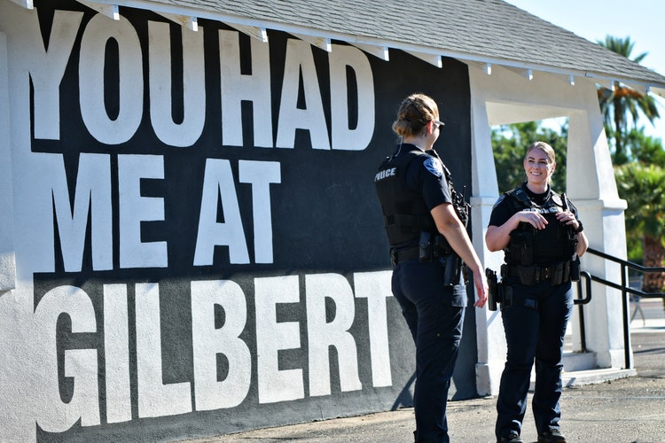 Two police officers in uniform stand smiling next to a building with a large mural that reads YOU HAD ME AT GILBERT. The scene is bright and sunny, with palm trees visible in the background.