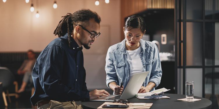 Two marketing executives work on an upselling campaign using a laptop at a table in a common area.