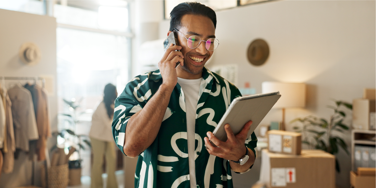 A man wearing glasses and a patterned shirt is smiling while talking on his phone and holding a tablet. He is standing in a bright, modern workspace with clothing racks, boxes, and plants visible in the background.