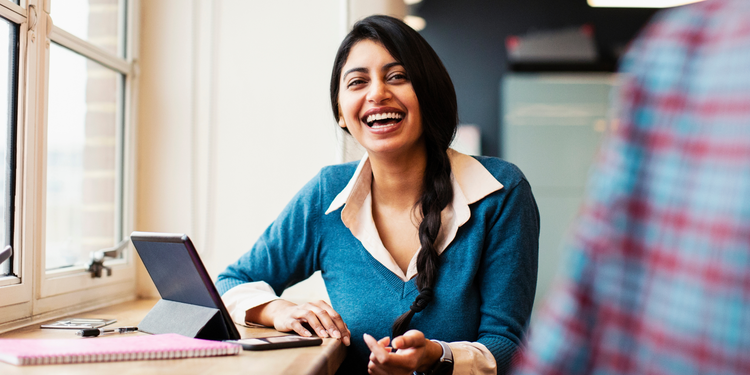 Smiling woman with a braid, wearing a blue sweater and white shirt, sitting by a window with a tablet, notebook, and pen on the table.