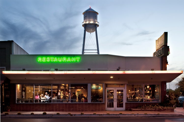 Exterior view of a restaurant at dusk, featuring bright neon signage reading RESTAURANT, a warmly lit interior with patrons dining, and a prominent water tower rising in the background.