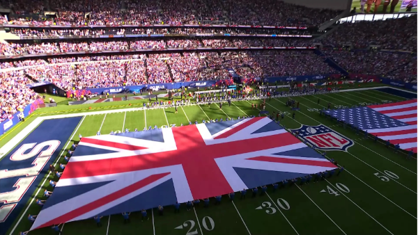 NFL stadium packed with fans as large Union Jack and U.S. flags are displayed on the field during a pre-game ceremony.