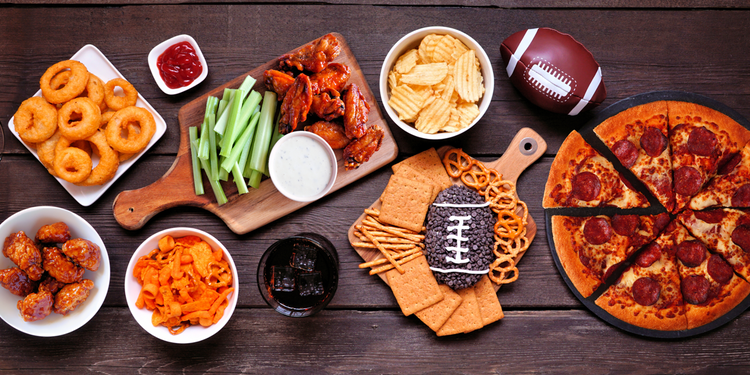 Super Bowl snacks displayed on a table — including onion rings, buffalo wings, potato chips, pretzels, and pizza.