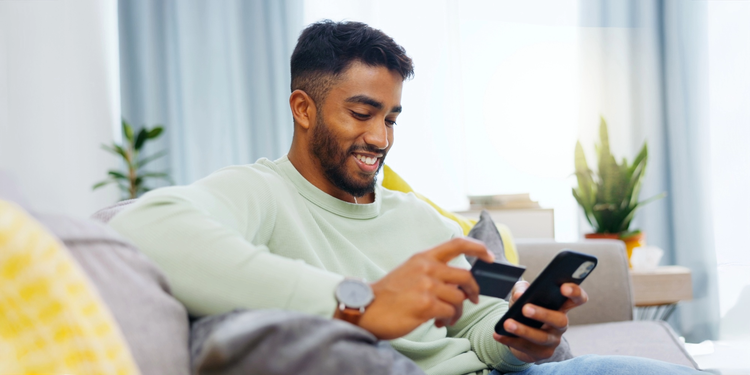 Man sitting on a couch, smiling while using a smartphone and holding a credit card.