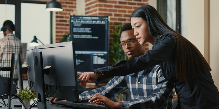 A woman with long black hair, wearing a black blouse, is leaning over and pointing at a computer screen while assisting a male colleague who is wearing glasses and a plaid shirt. They are both focused on the screen, which displays lines of code.