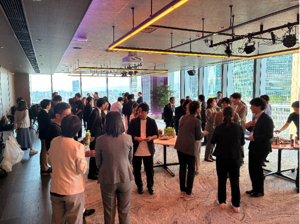 The image depicts a lively and crowded indoor event with people mingling and participating, under a modern, well-lit, high-ceilinged, and glass-walled setting with an urban skyline visible through large windows. AI 生成コンテンツは誤りを含む可能性があります。