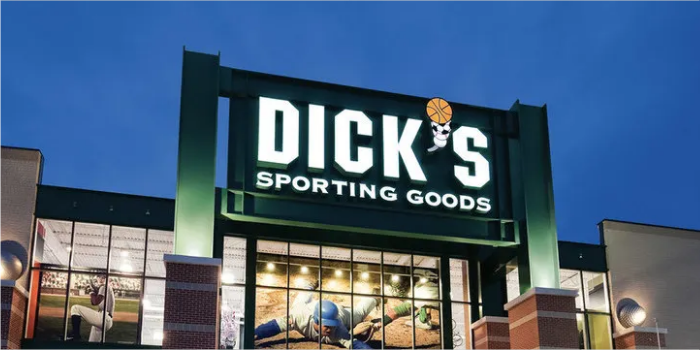 Exterior of Dick's Sporting Goods store at dusk, with large illuminated sign and sports-themed window display below.