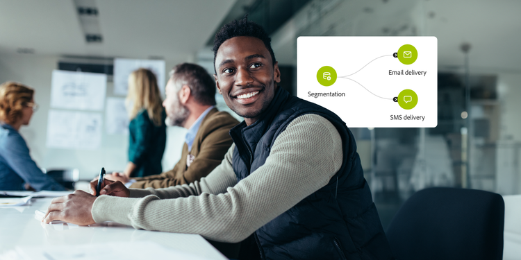 A smiling man sits at a conference table with colleagues in a bright, modern office setting. A graphic beside him illustrates audience segmentation branching into two paths labelled email delivery and SMS delivery.