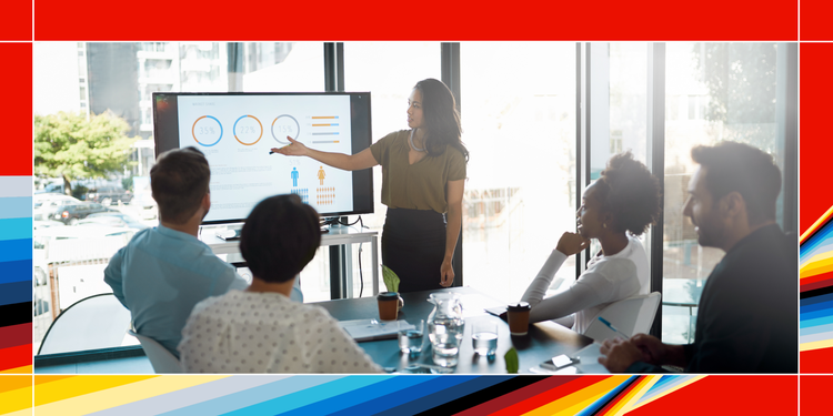 In a corporate environment, an instructor standing in front of a desk and a screen speaks to a small group of employees sitting in front of her.