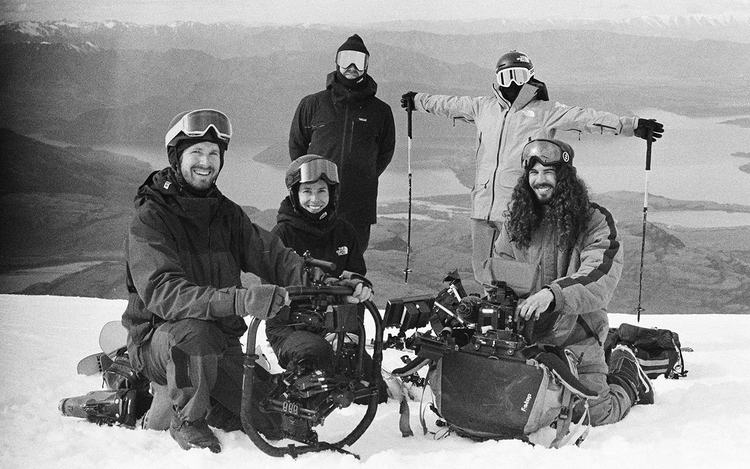 Black and white picture of five people in winter gear on a snowy mountain with camera equipment and ski poles, with a lake behind.