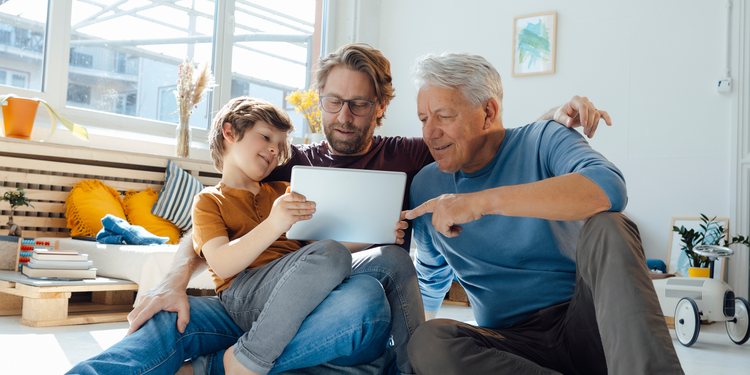 A grandfather, a father and a son looking at a tablet sitting in a bright family room.