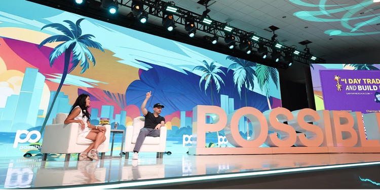 A stage at a conference featuring two speakers seated in white chairs, engaging in discussion. A colourful, tropical-themed backdrop with palm trees and cityscape imagery fills the screen behind them. Large, bold letters spelling 'POSSIBLE' stand prominently on the stage, with a presentation slide reading ‘I day trade attention and build businesses.’