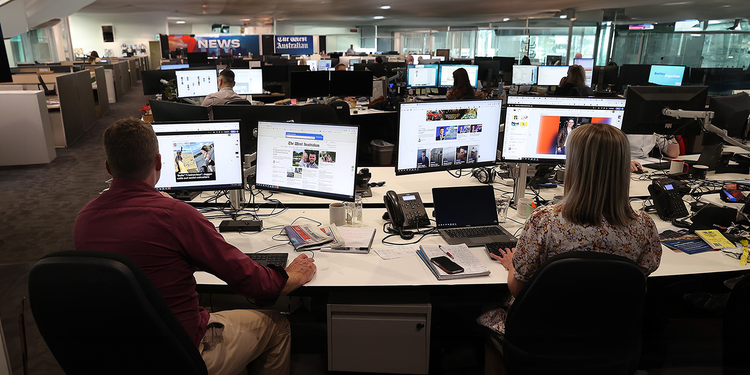 Image of two people sitting at a desk working on computers — within a larger office full of other people.
