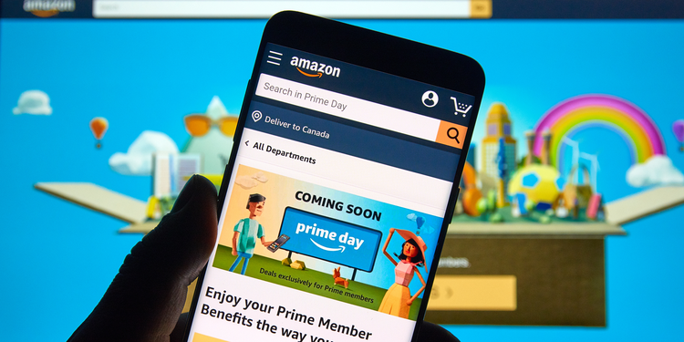 Person holding a smartphone displaying the Amazon app with a Prime Day promotion, against a colourful Prime Day-themed background on a monitor.