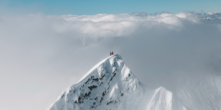 Two climbers standing on a snow-covered mountain peak, with clouds below and clear sky above.