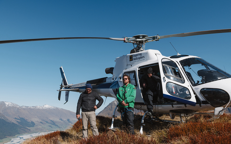 Three people in outdoor gear stand beside a helicopter named “Aspiring Helicopters” on a grassy hilltop with mountains in the background.