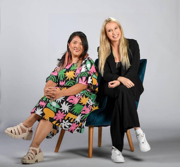 Image of two women seated on a blue chair. One is wearing a bright patterned dress, the other is wearing a dark suit.