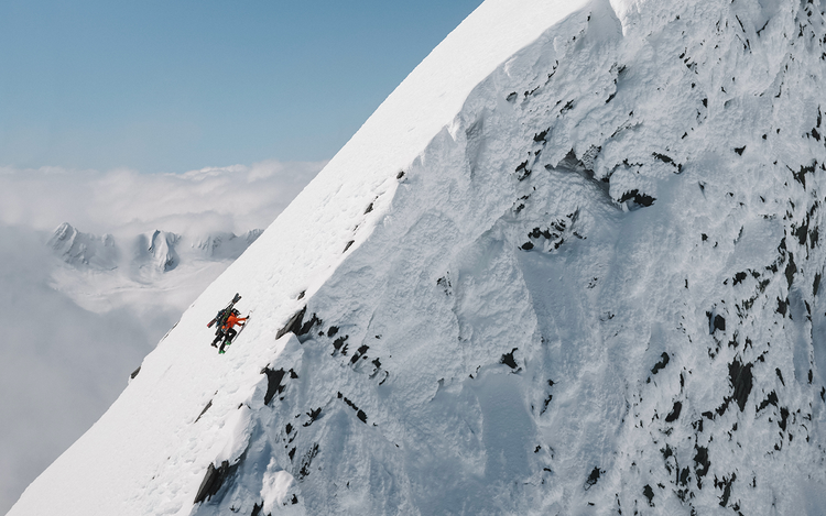 Two climbers ascending a steep, snow-covered mountain slope with gear and ice axes, set against a clear sky.