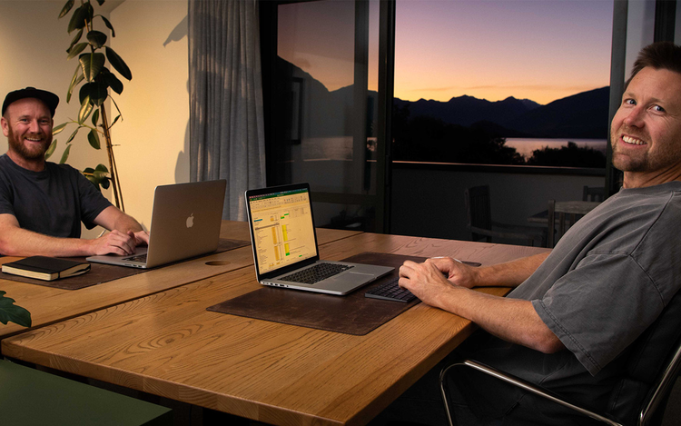 The two founders working on laptops at a wooden table with a mountain sunset in the background, showcasing remote work and collaboration.