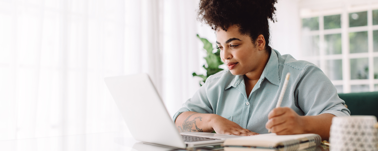 A female business professional writes on a notebook and uses a laptop to manage projects with Adobe Workfront.